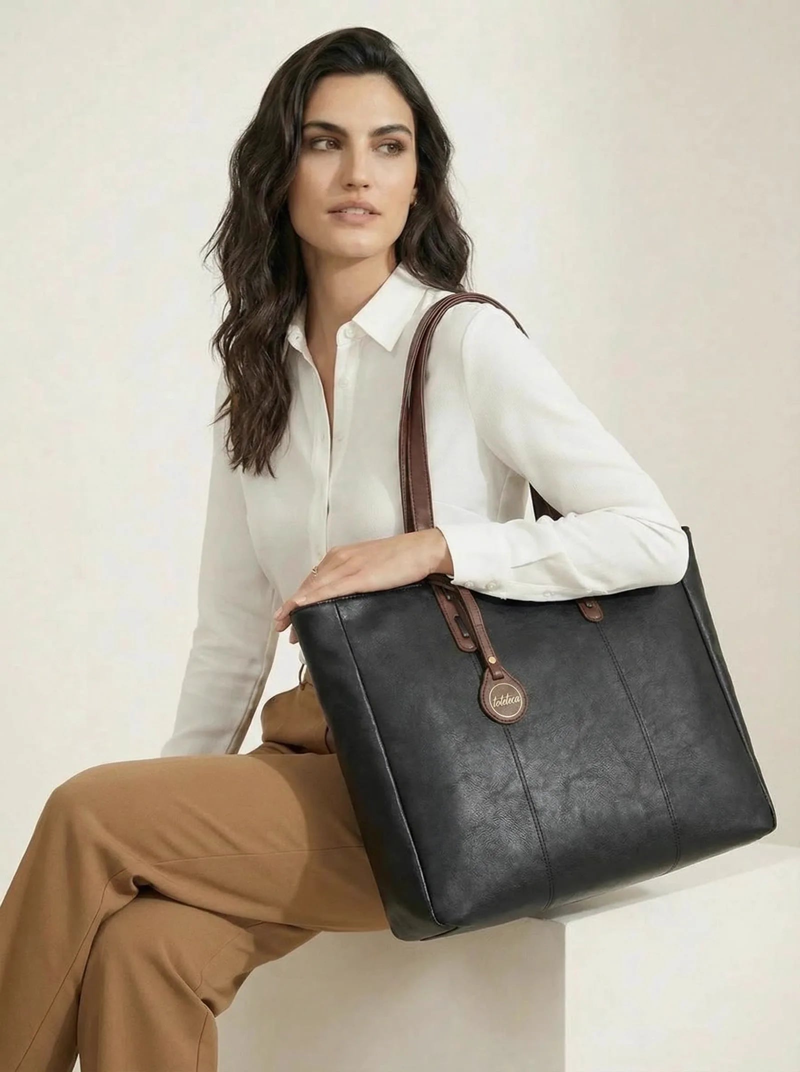 Model seated with a black Toteteca tote with brown handles and logo charm